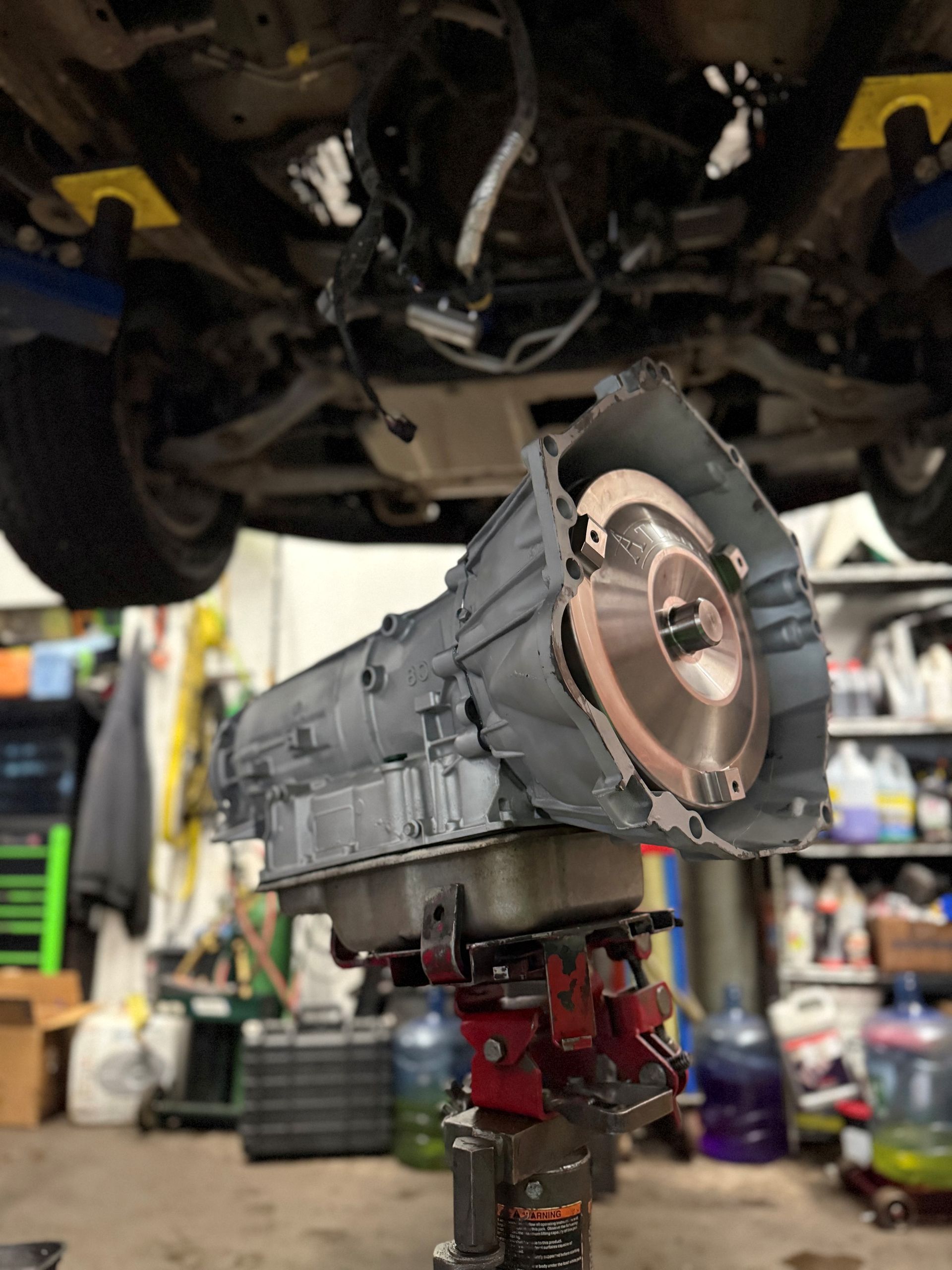 A car transmission, light gray, sits on a lift below a vehicle in a garage setting.