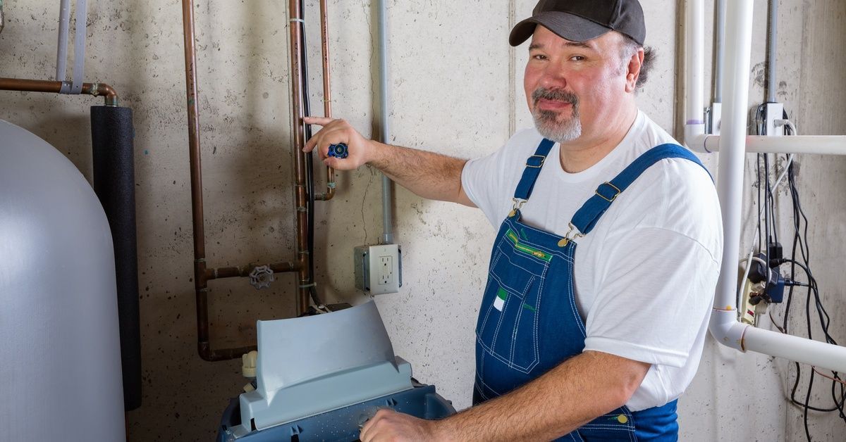 A man in overalls holds a wrench and stands beside an open water softener. His free hand is on a val