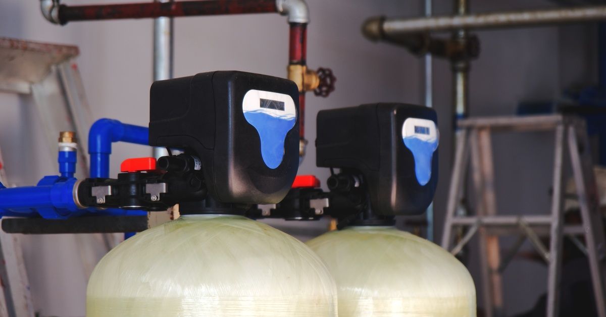 Two water softener tanks with black control heads. There are numerous pipes and ladders behind them.