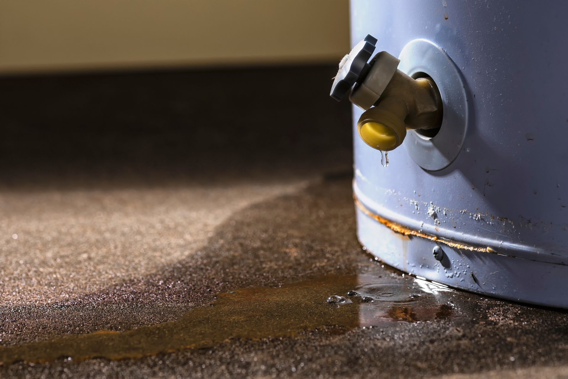 A leaking water heater with water dripping from a spout onto a small puddle on the concrete floor.