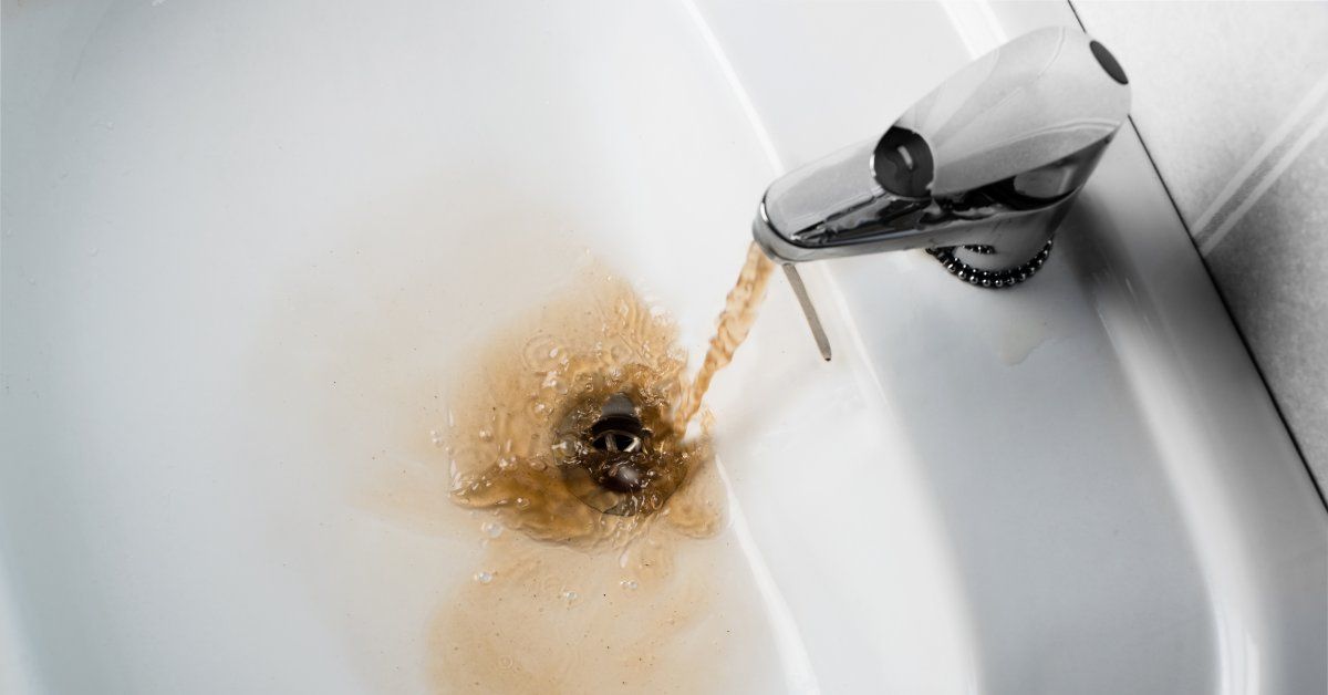 A faucet on a white sink pouring discolored water. The water pools in the sink before flowing down.