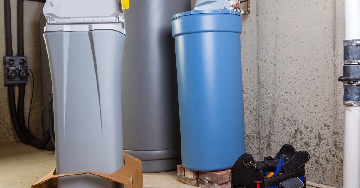 A water softener system standing on bricks in a basement. A new system still in its packaging stands nearby.