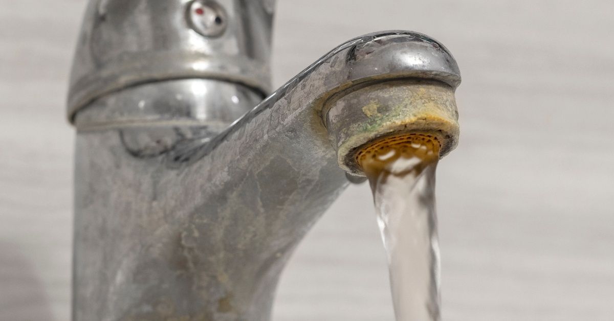 A close-up of a sink faucet covered in hard water stains and other grime. Water is pouring out of the faucet.
