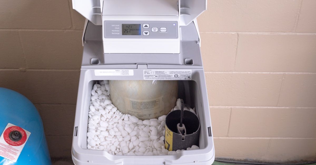 A water softener with the lid open positioned against a stone wall. The tank is full of salt pellets
