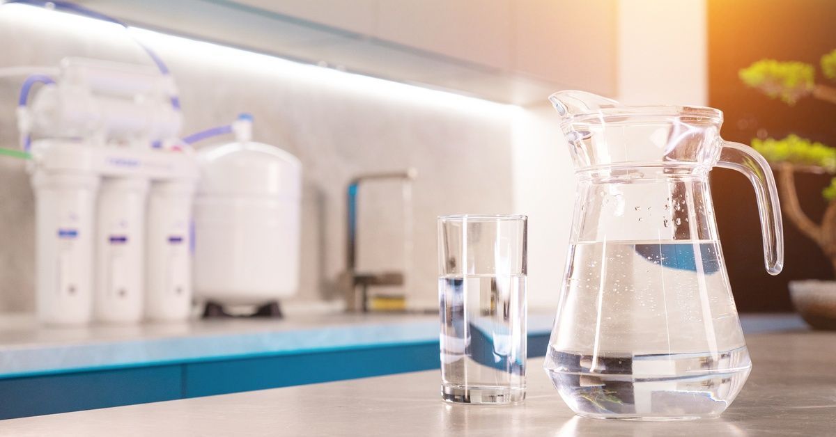 A glass pitcher and cup full of clean, clear water. Behind them is a reverse osmosis water filtratio