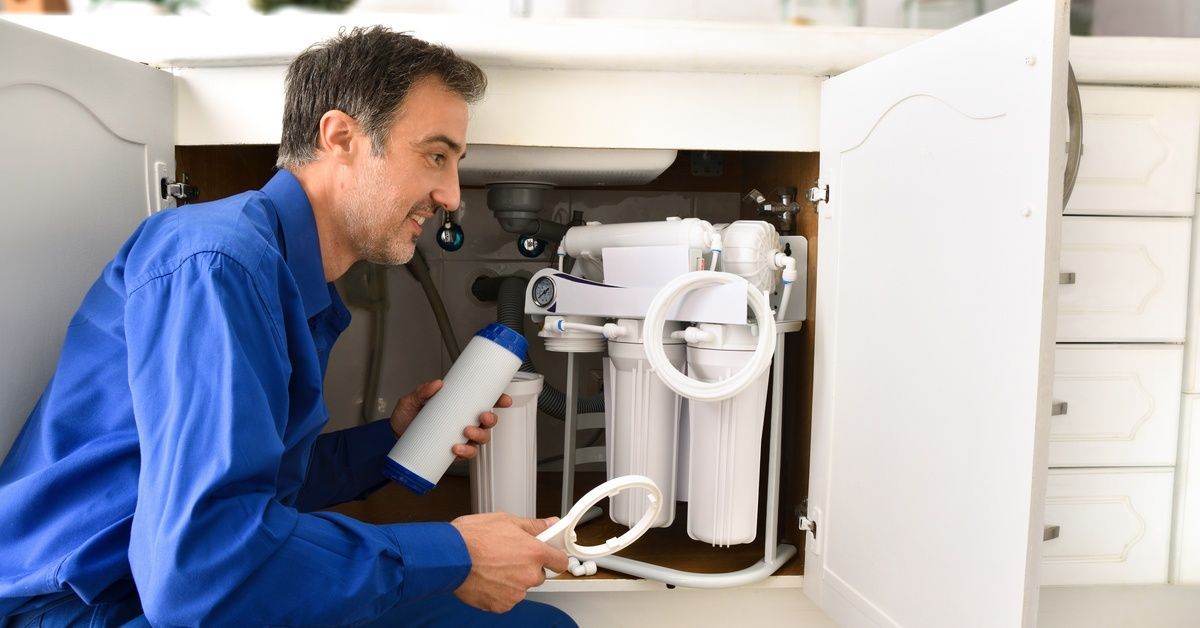 A technician in a blue shirt kneeling in front of a cupboard beneath a sink and working on a reverse osmosis system.