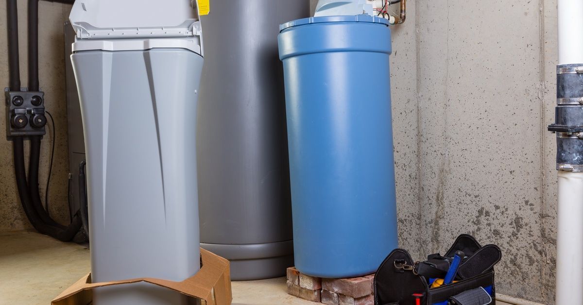 A new water softening unit still in a cardboard holder standing beside an old unit and a bag of tools.