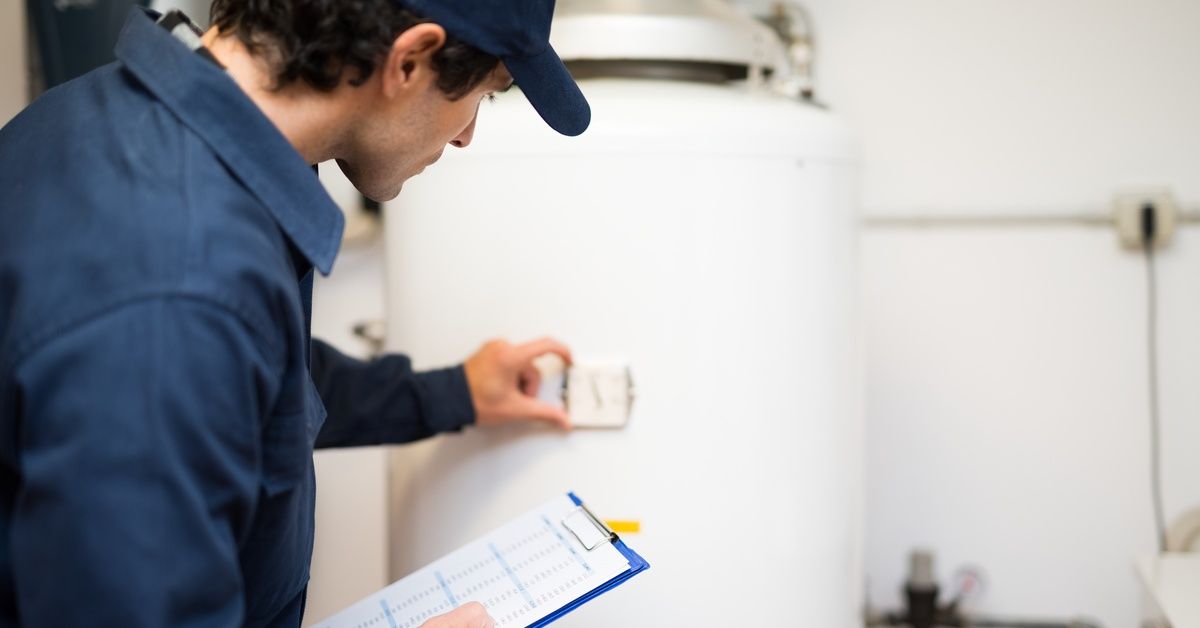 A technician in a work shirt and hat holds a clipboard in one hand and inspects a water heater tank.