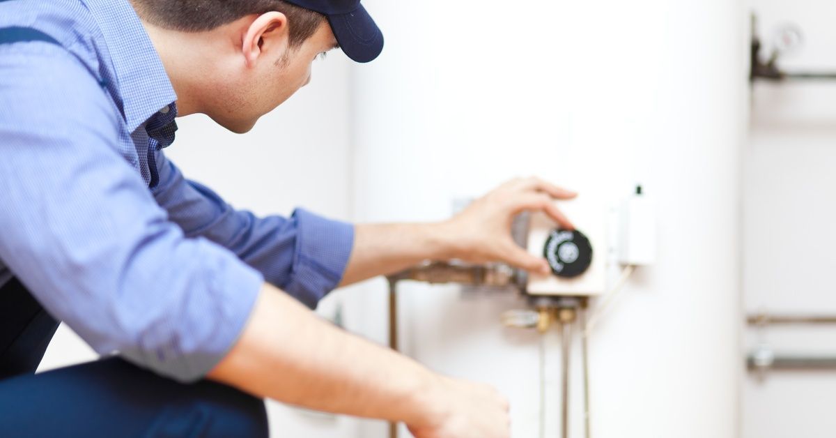 A worker in a blue shirt, pants, and hat kneels beside a water heater tank and touches a dial on it.