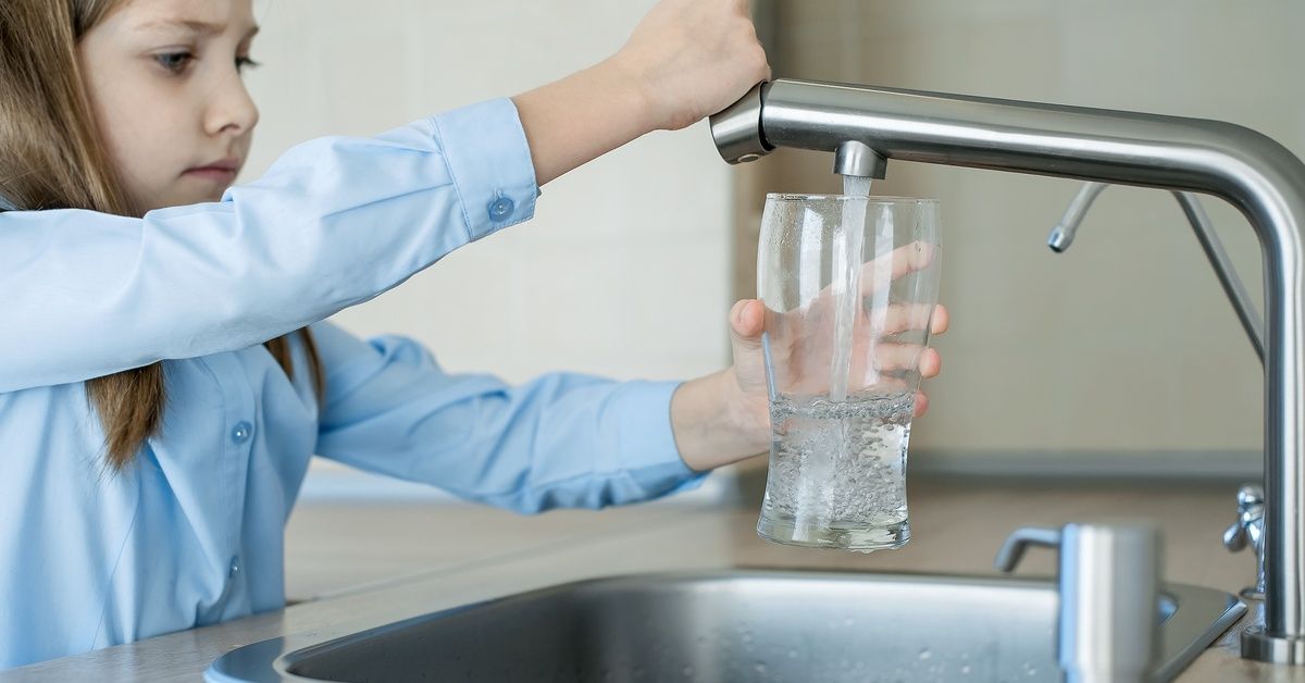 A little girl standing at a sink and filling a drinking glass with water from the faucet.