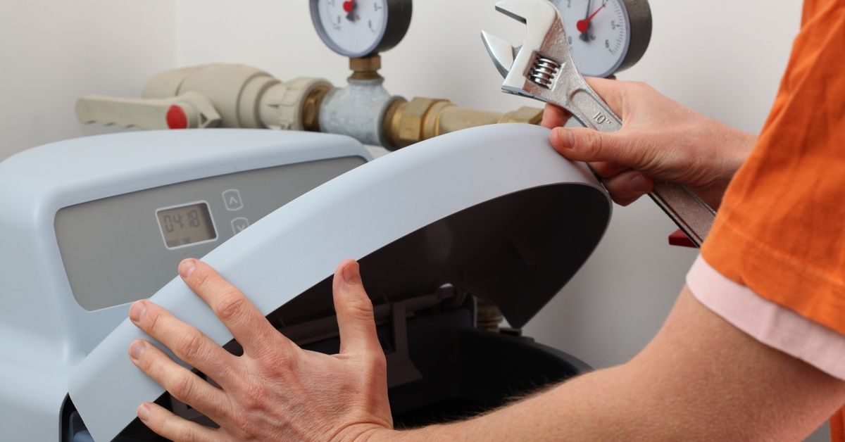 A person wearing an orange shirt and holding a wrench stands next to an open water softener.