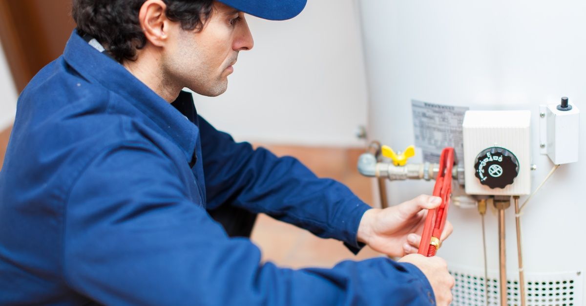 A plumber in a blue uniform kneeling in front of a water heater, using a wrench to unscrew a pipe.