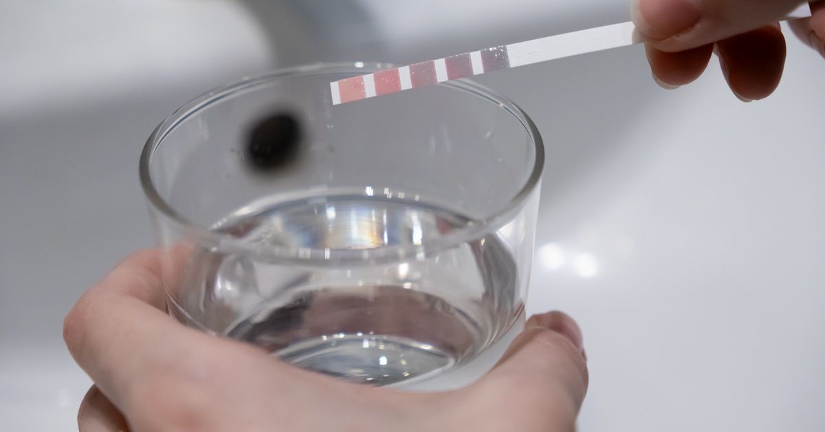 A person holding a glass of water, testing it with a strip turning red/purple in their other hand.