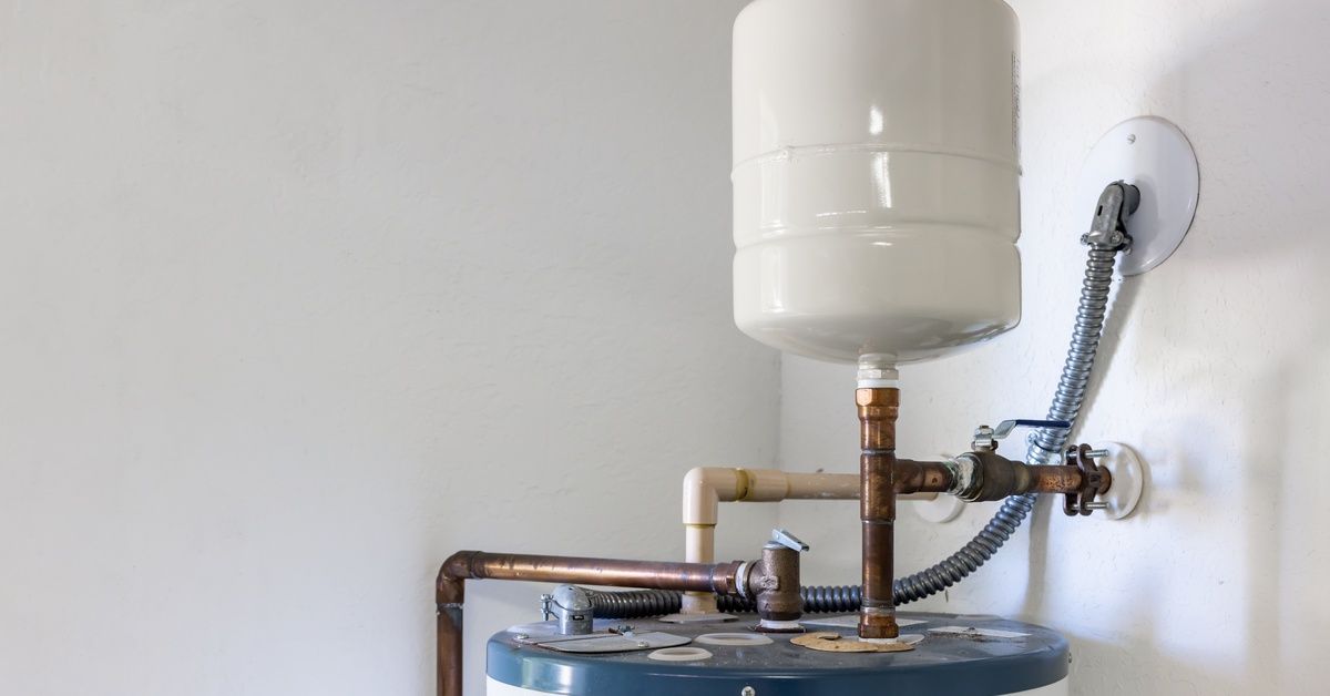 A white tank attached to a larger machine with brown pipes, and to the wall by a ridged steel pipe.