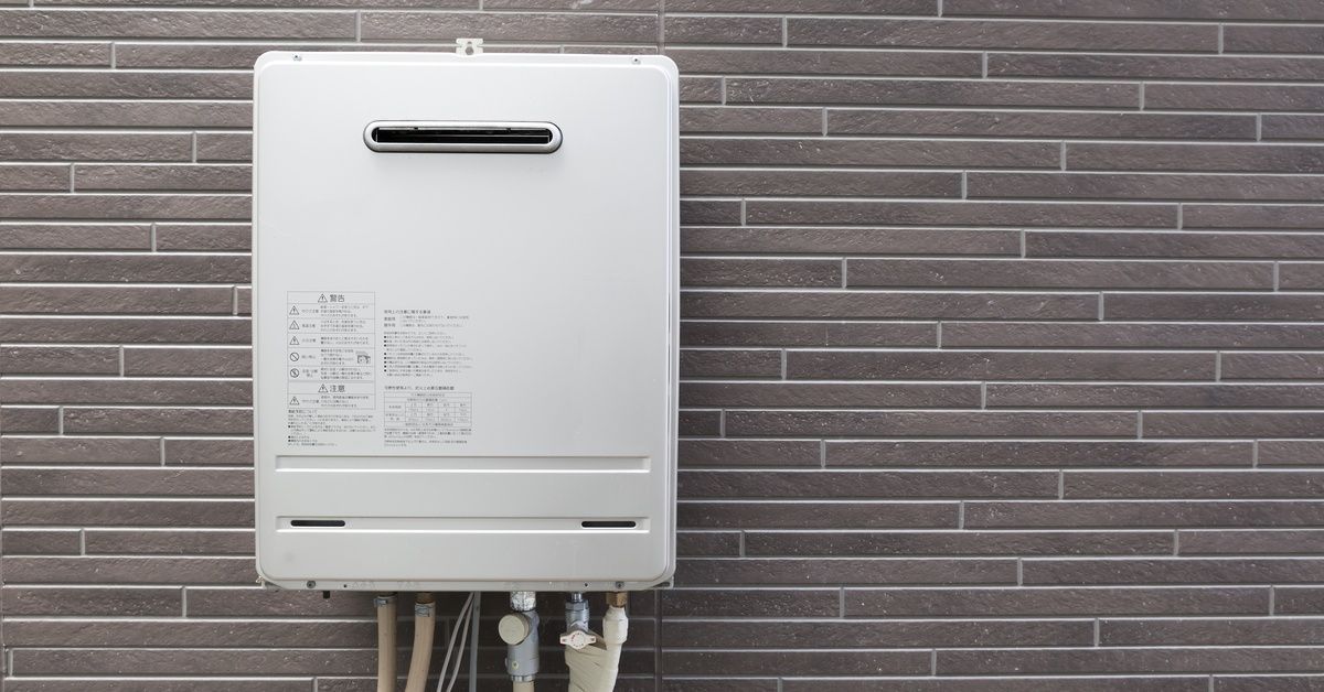 A boxy white water heater connected to several thin hoses against a brown-gray brick wall.