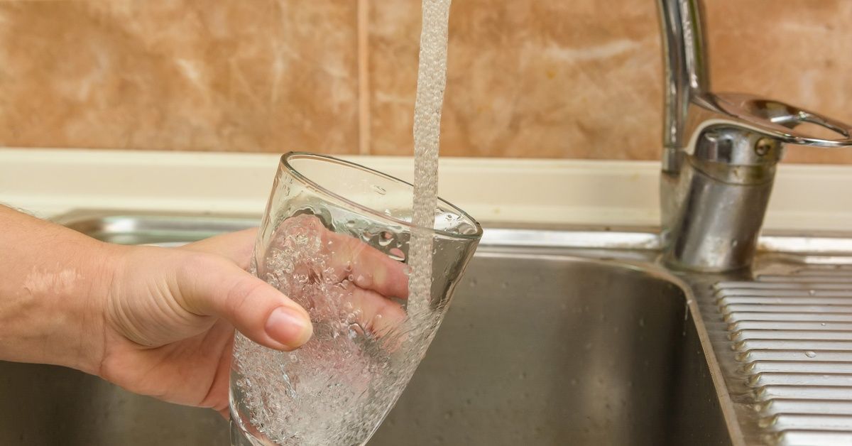 A hand holding a clear glass cup directly under a sink faucet as a steady stream of water pours and splashes inside.