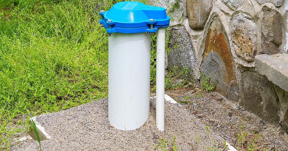 A white and blue water well casing near a stone wall, surrounded by gravel neatly spread at its base