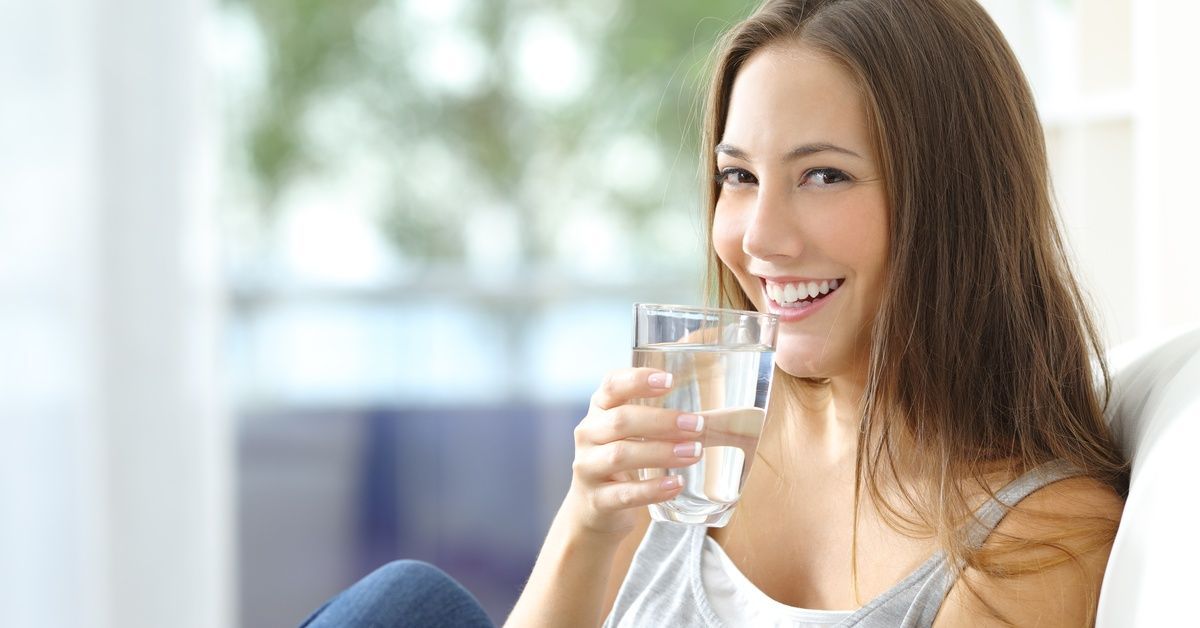 A smiling woman sitting on a couch by the window with sunshine streaming in. She holds a glass of water up to her lips.