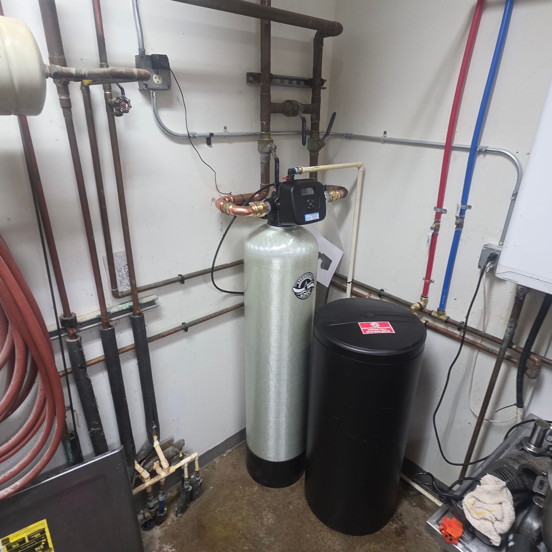 Utility room with water softener tanks, pipes, and hoses along the wall