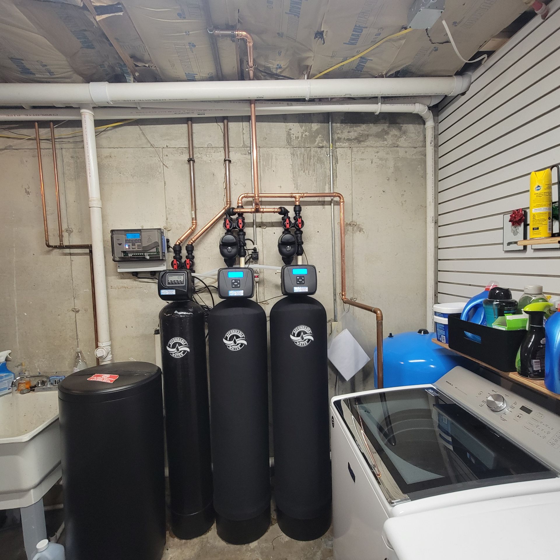Basement utility area with water treatment tanks, pipes, and a washing machine under exposed ceiling pipes