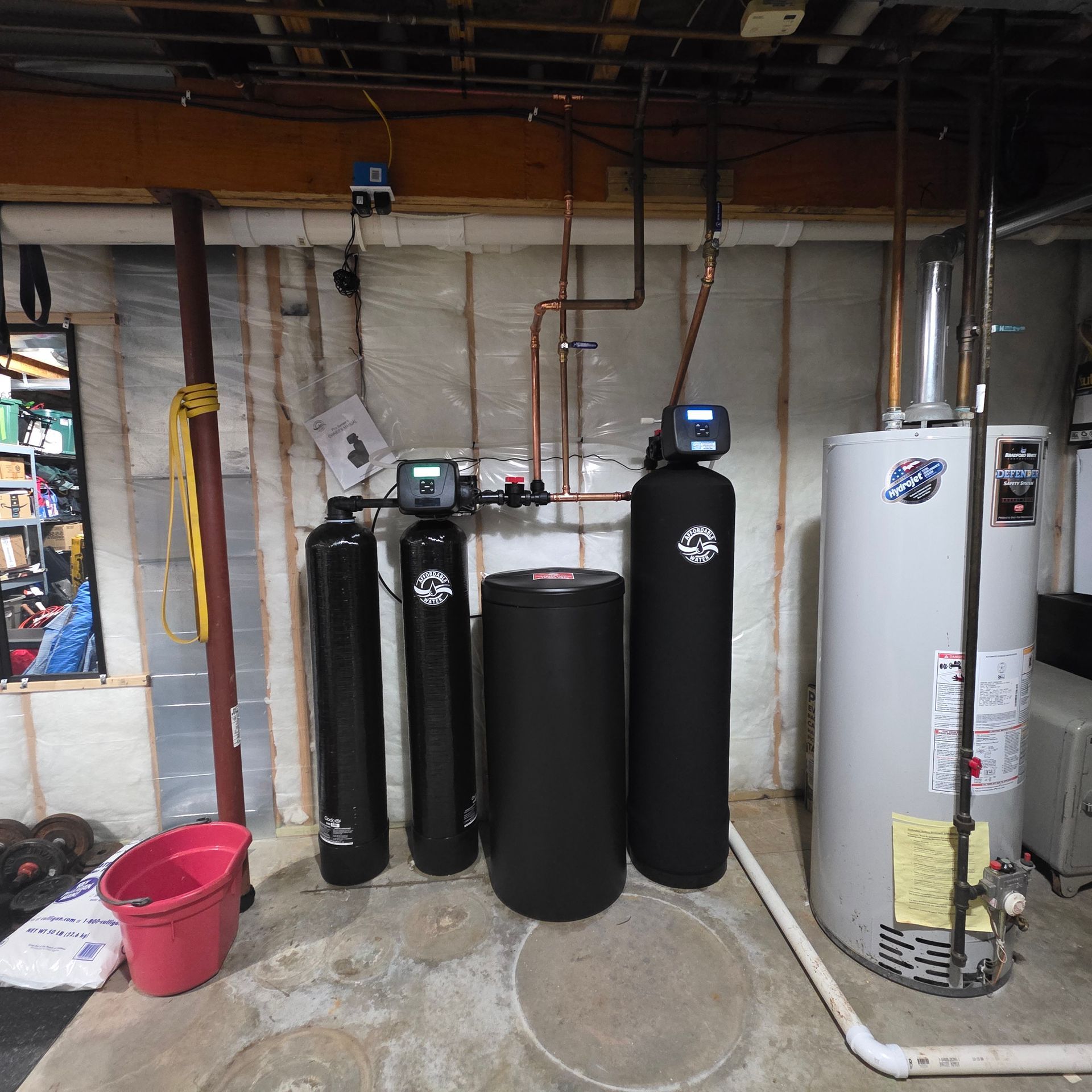 Basement utility room with water softeners, tanks, pipes, and a red bucket on the concrete floor