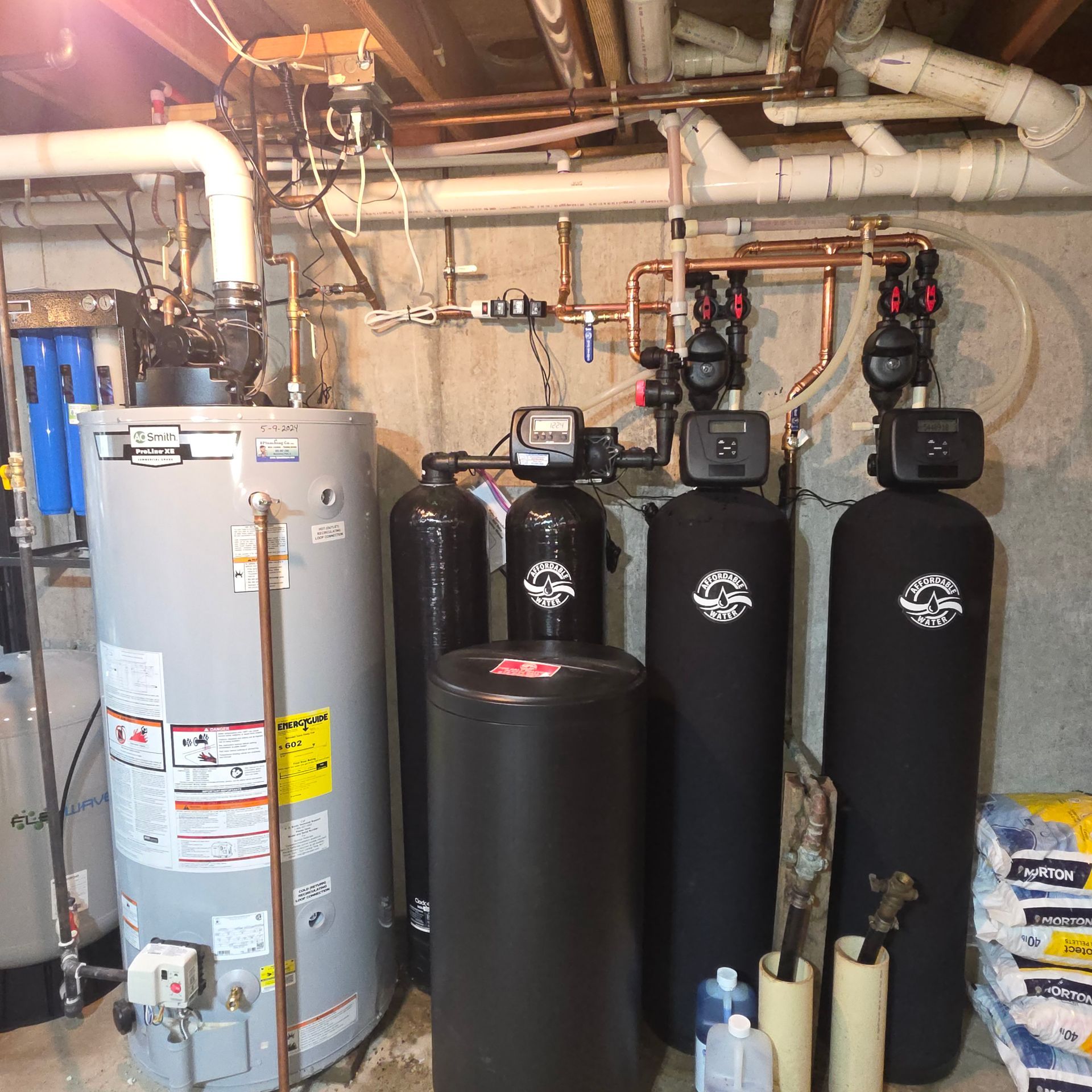 Basement water treatment system with large gray tank, black filters, and exposed pipes and valves