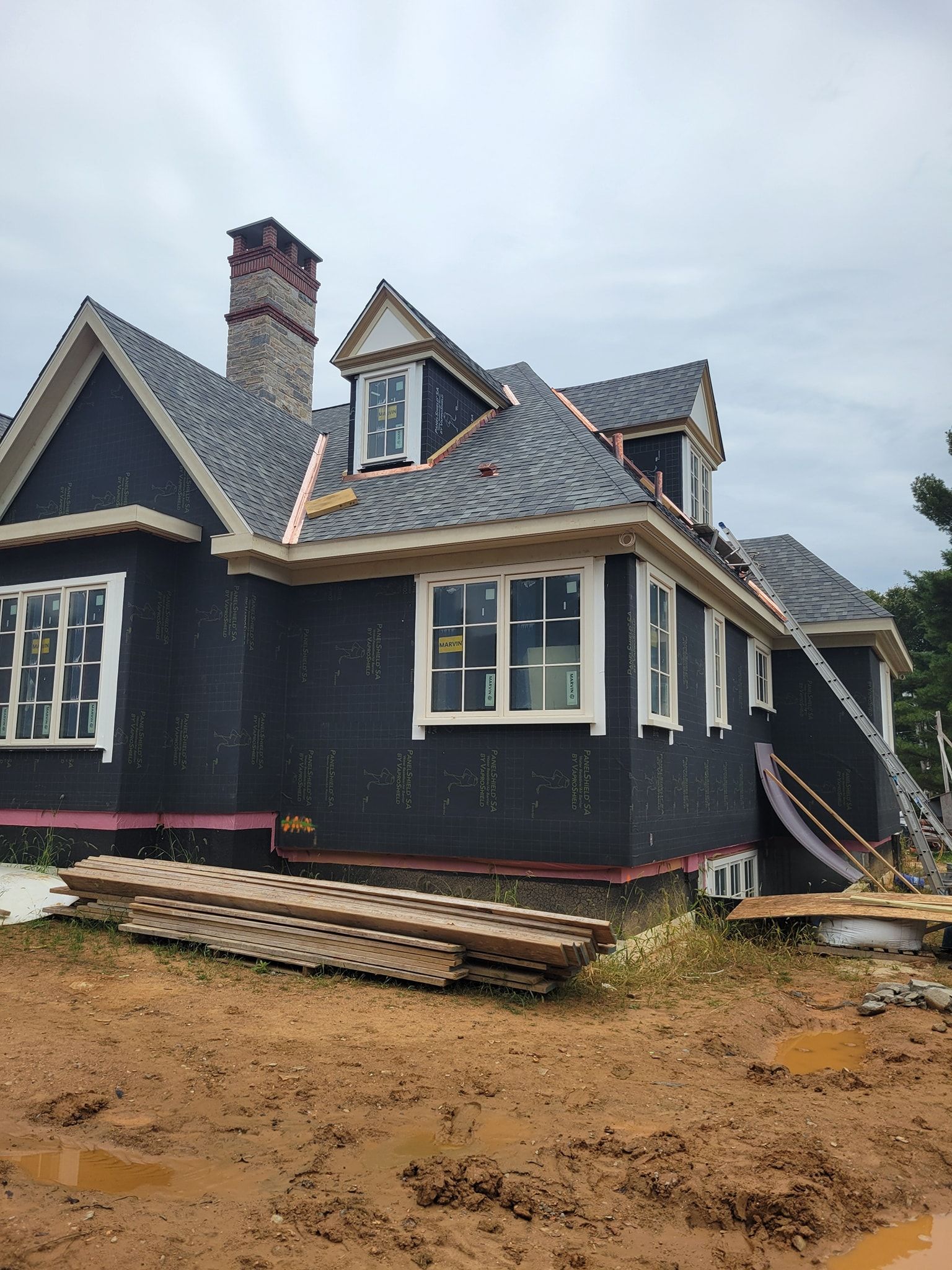 House under construction, dark siding, white trim, grey roof, chimney, windows, brown muddy ground, boards.