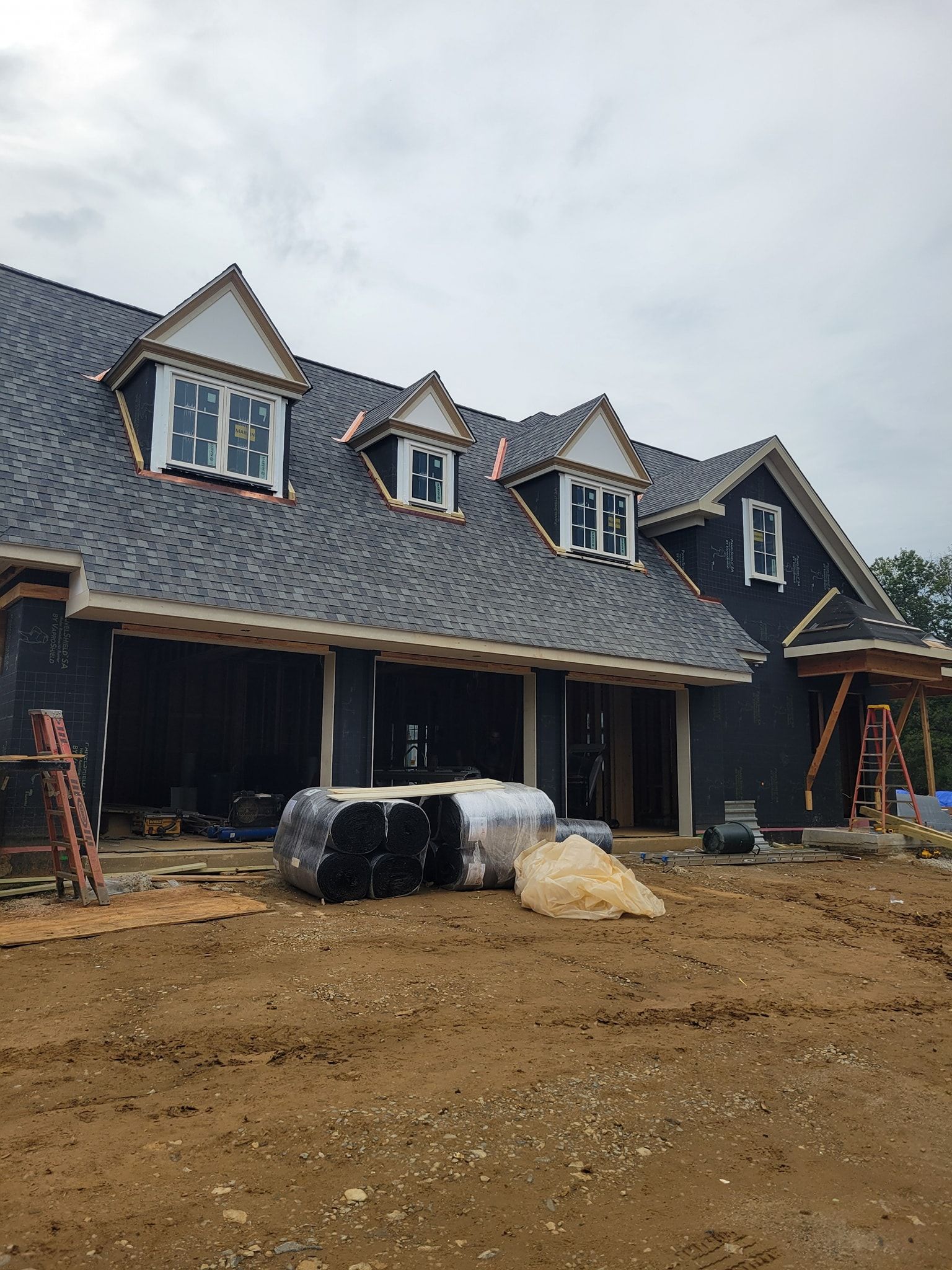 House under construction; black sheathing, grey shingles, three dormers, brown soil.