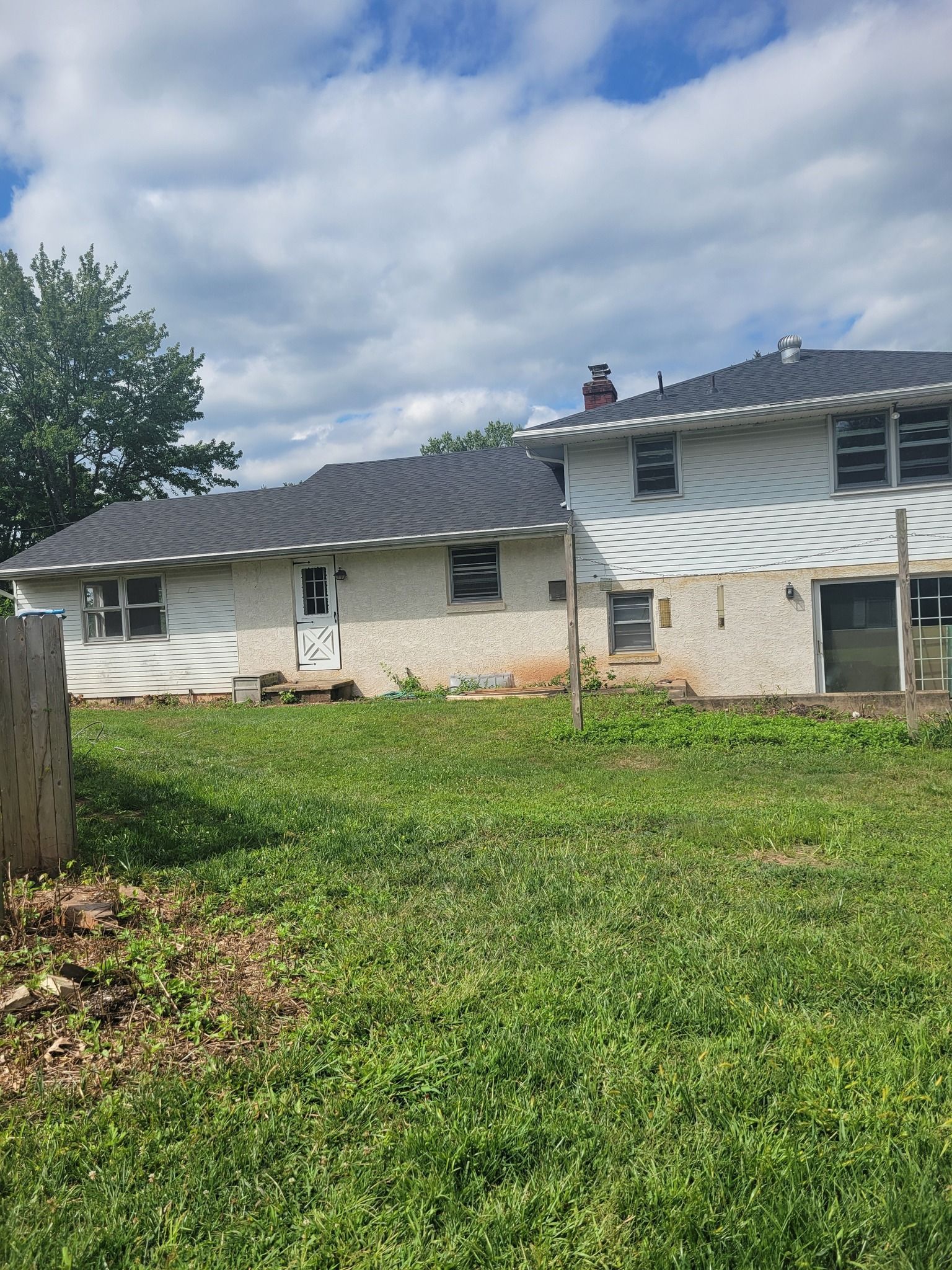 Backyard view of a two-story white house with a dark roof and chimney, set against a cloudy blue sky.
