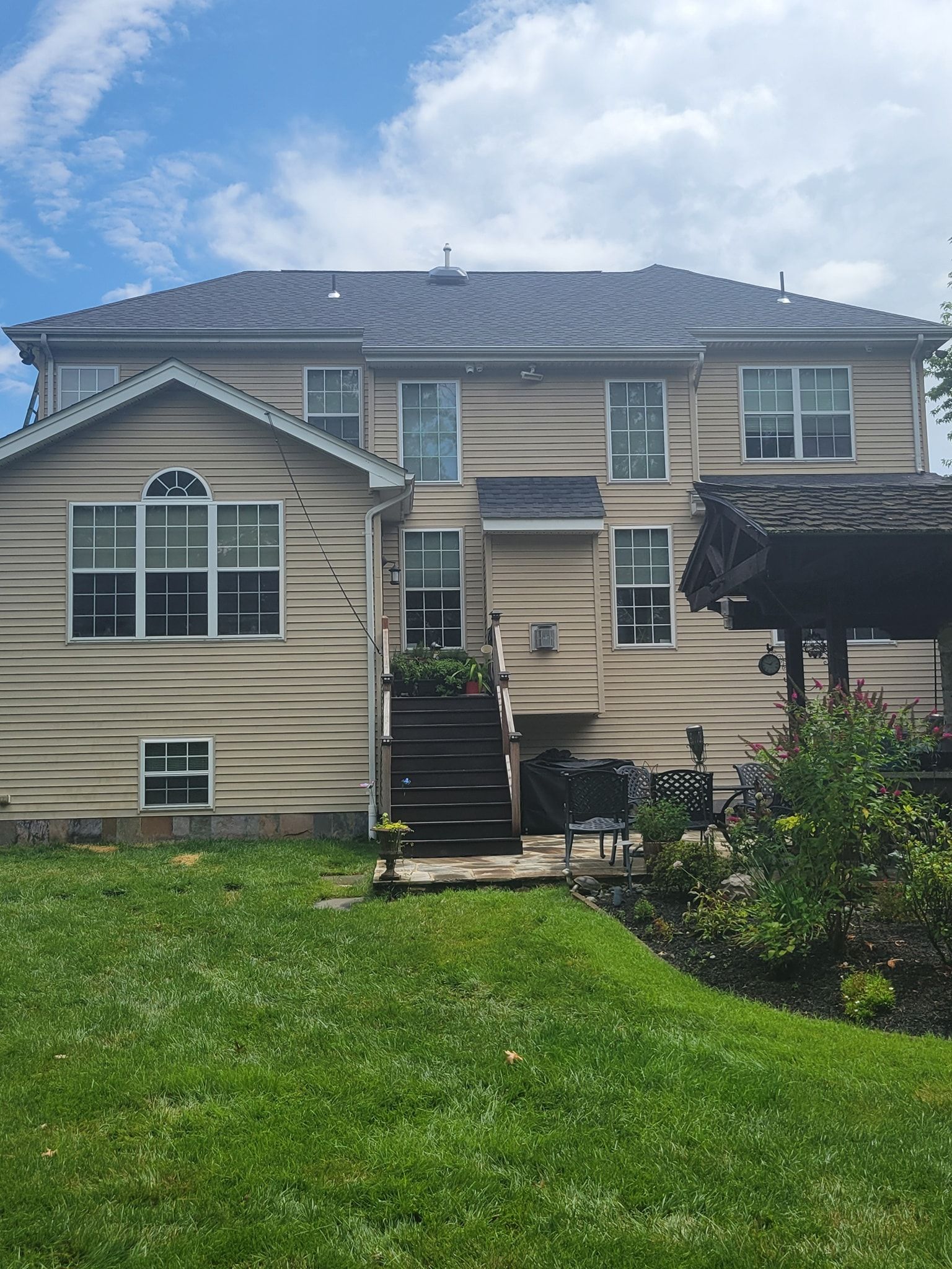 Back of a two-story beige house with a dark roof, stairs, windows, and green lawn.