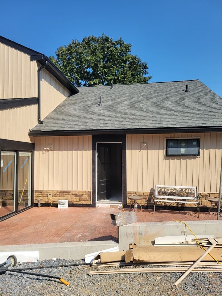 Exterior of building with tan siding, black trim, and a brick patio under construction.