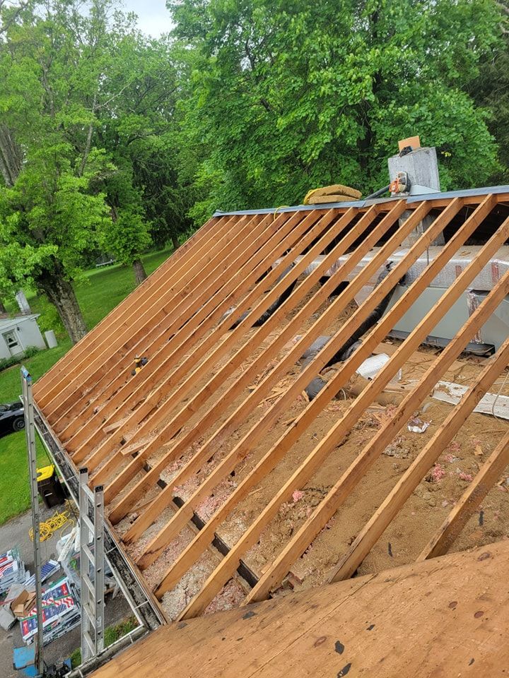 Roof under construction with exposed wooden beams, surrounded by green trees.