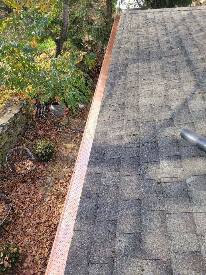 Copper rain gutter along the edge of a gray asphalt shingle roof. Autumn leaves on the ground.