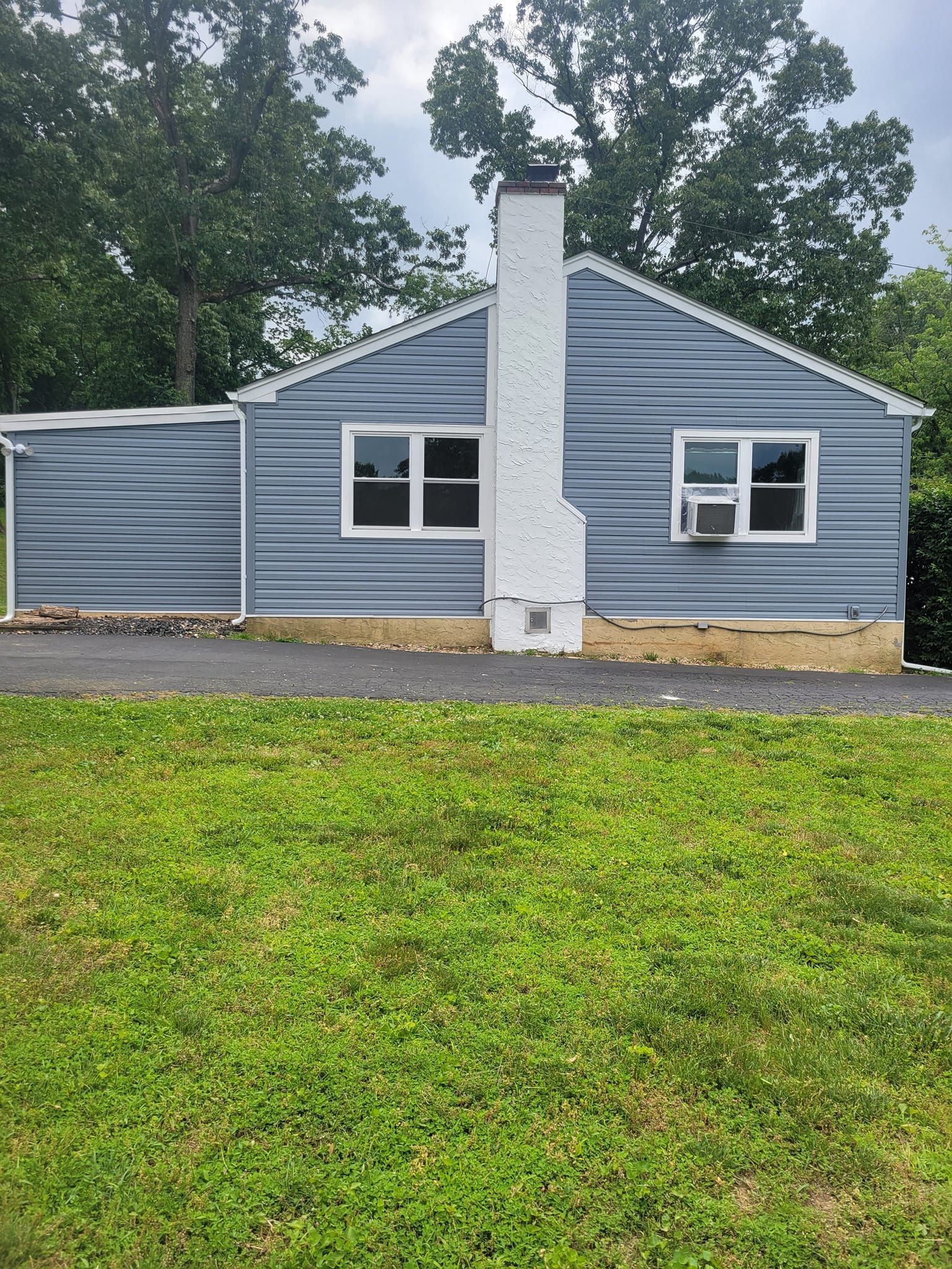 Blue-sided house with a white chimney, two windows, and an air conditioner, in front of green grass.
