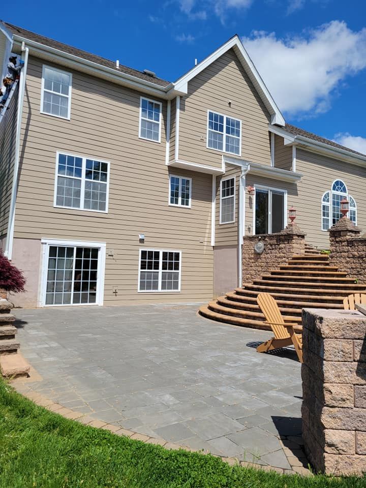 Rear exterior of a house with a stone patio, steps, and tan siding. Sunny day with a chair.