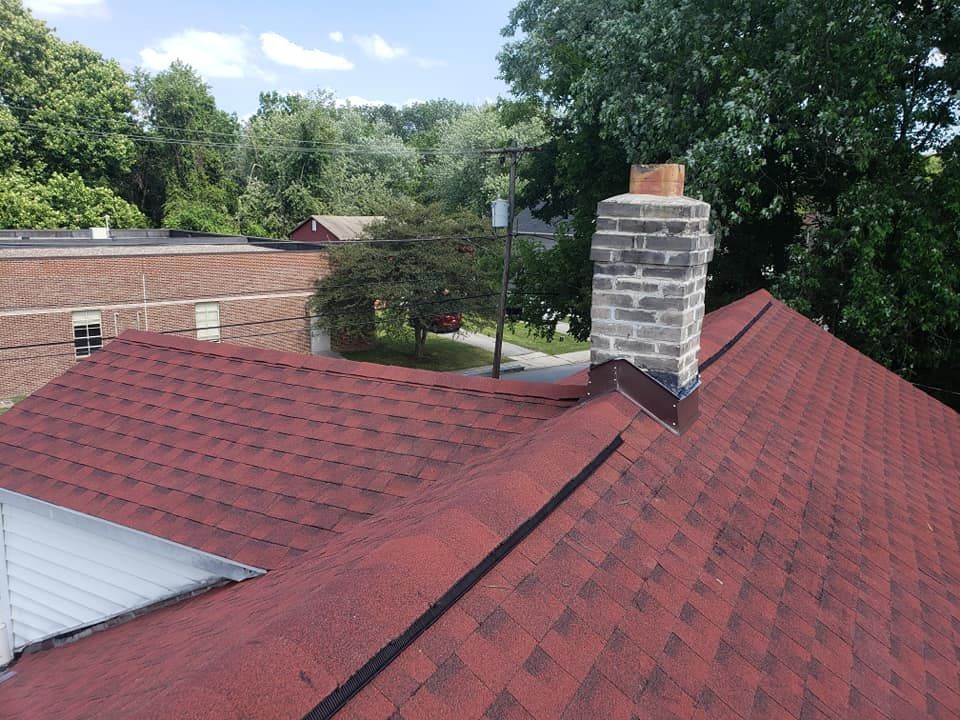 Red shingled rooftop with a brick chimney against a backdrop of trees and other buildings.
