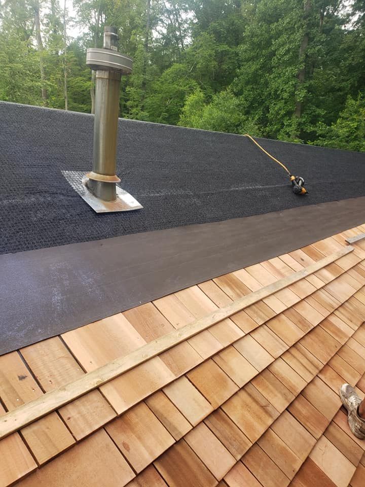 A roof under construction. Cedar shingles on the bottom, transitioning to black roofing material near a metal chimney.