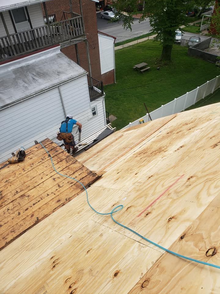 Roofer on a residential roof, installing new plywood. Beige wood, blue rope, white building, green lawn.