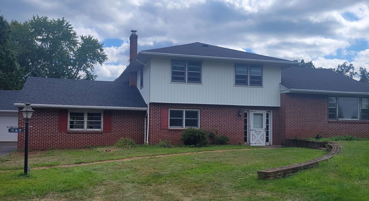 Two-story brick house with a dark roof and overgrown lawn under a cloudy sky.