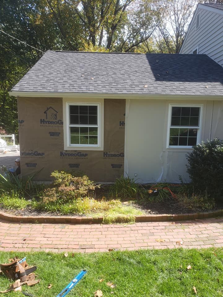 Exterior of a building under construction, showing a section with new sheathing and another with stucco. Two windows.