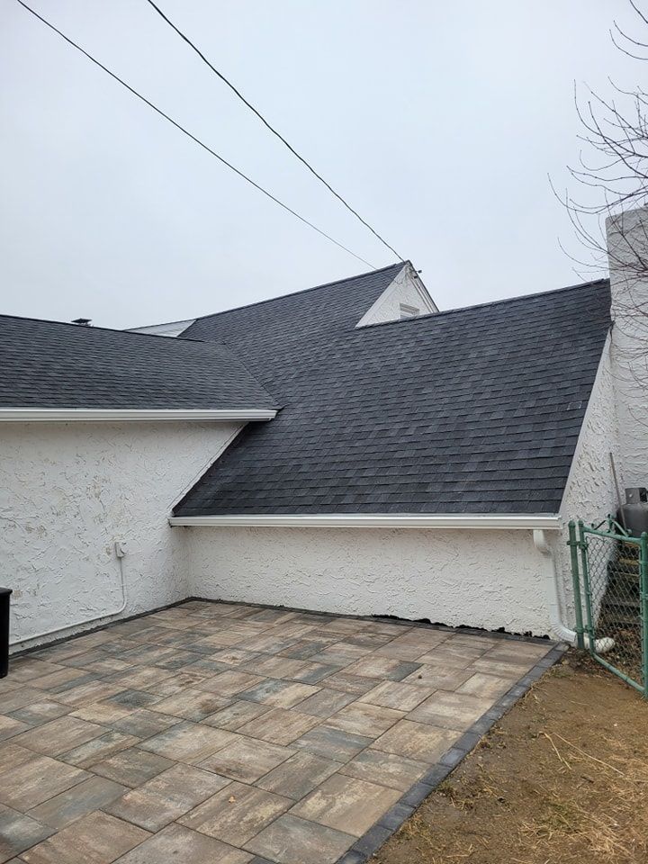White stucco house with dark gray shingled roof, patio pavers, and a chain link fence. Overcast sky.