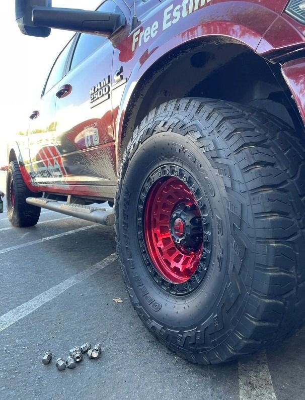 Red-rimmed truck tire on asphalt, with a red truck and side step. Gray lug nuts on the ground.