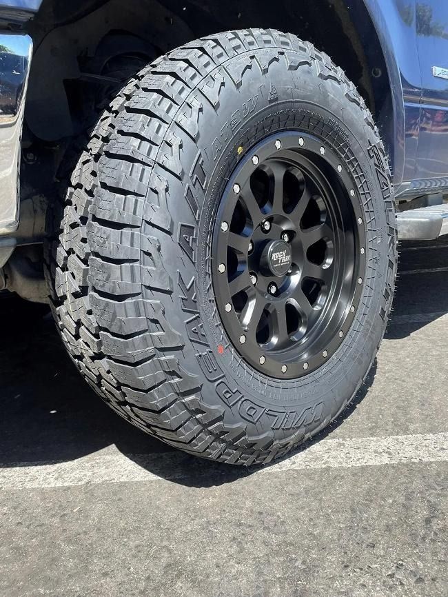 Black off-road tire on a black rim, mounted on a truck, parked on pavement.