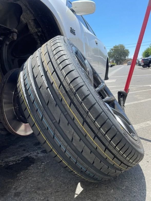 A car tire being replaced; a new tire in foreground, car raised with jack on a sunny day.