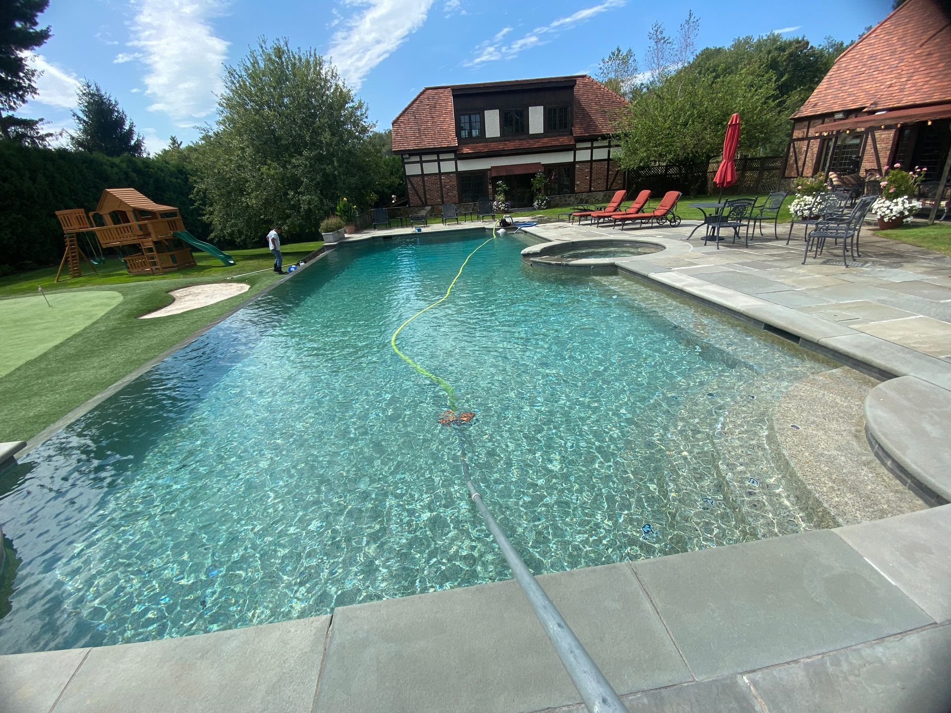 A rectangular pool with clear water, stone edges, and a house in the background under a blue sky