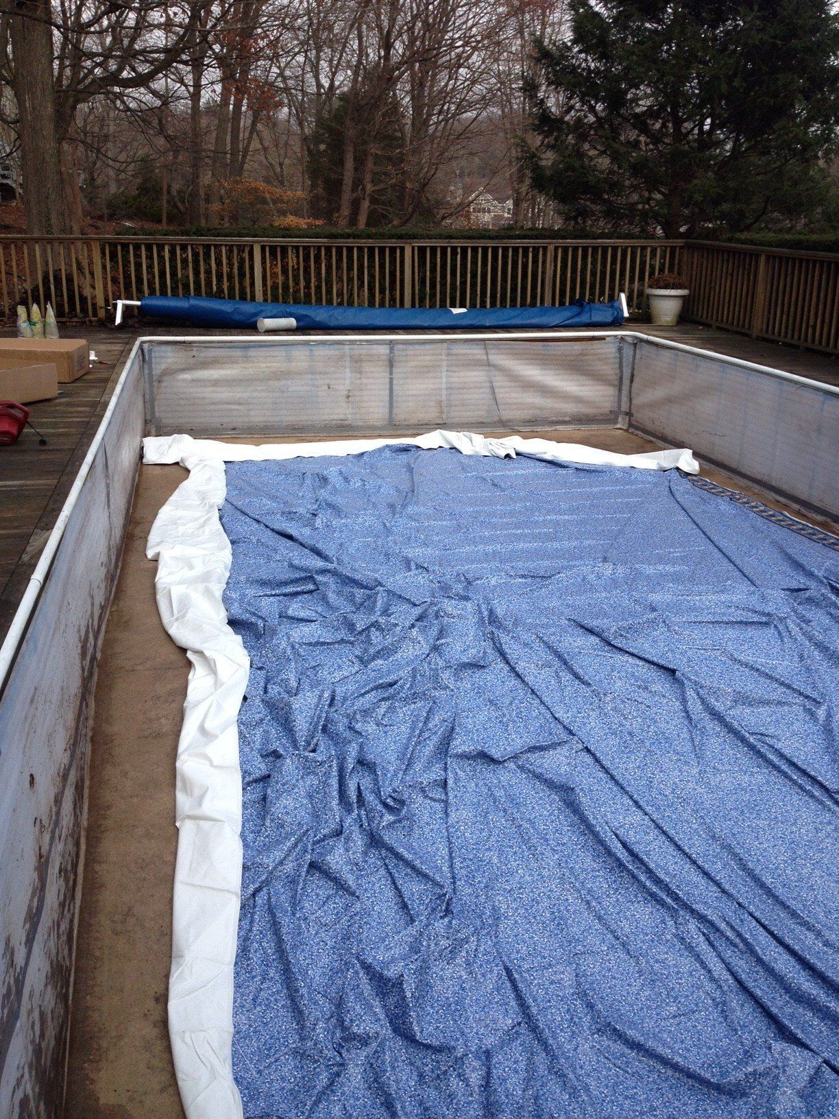 A rectangular, empty pool with a partially unfolded blue cover on a wooden deck.