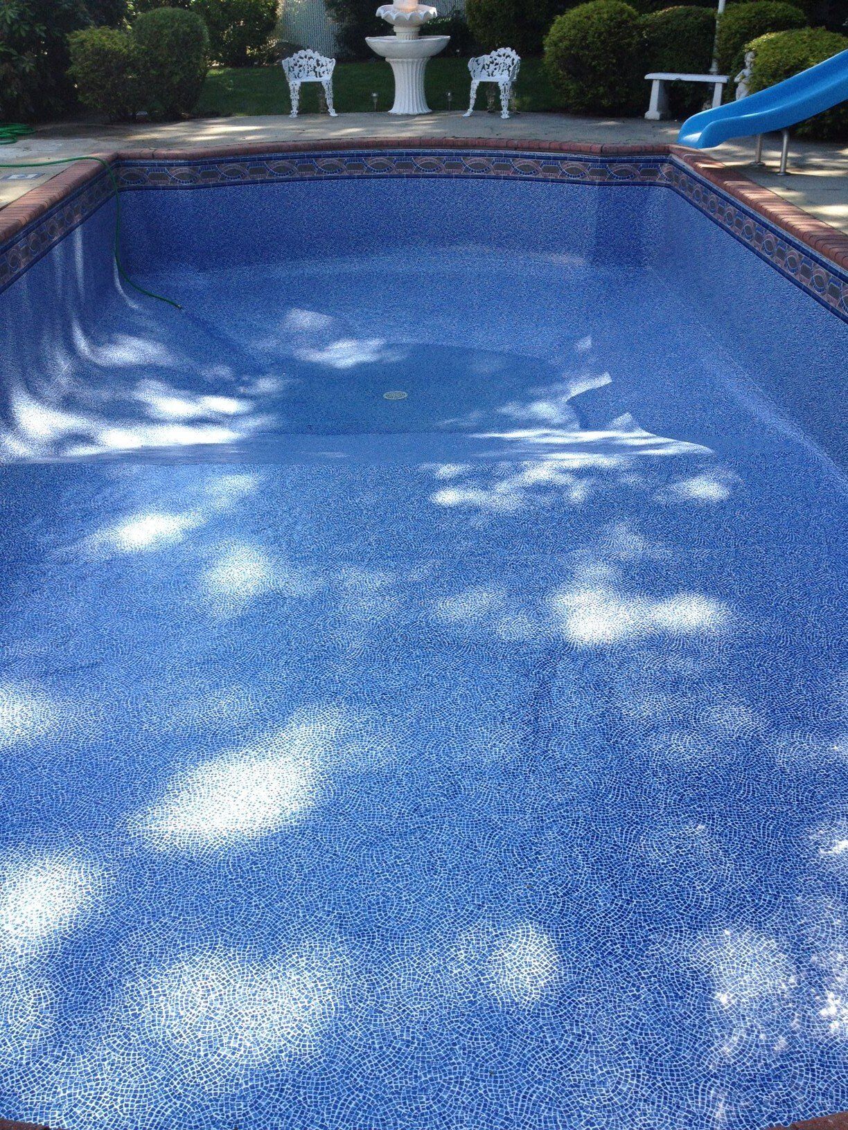 Blue mosaic-tiled swimming pool reflecting sunlight, set in a brick-lined patio with a fountain and garden visible