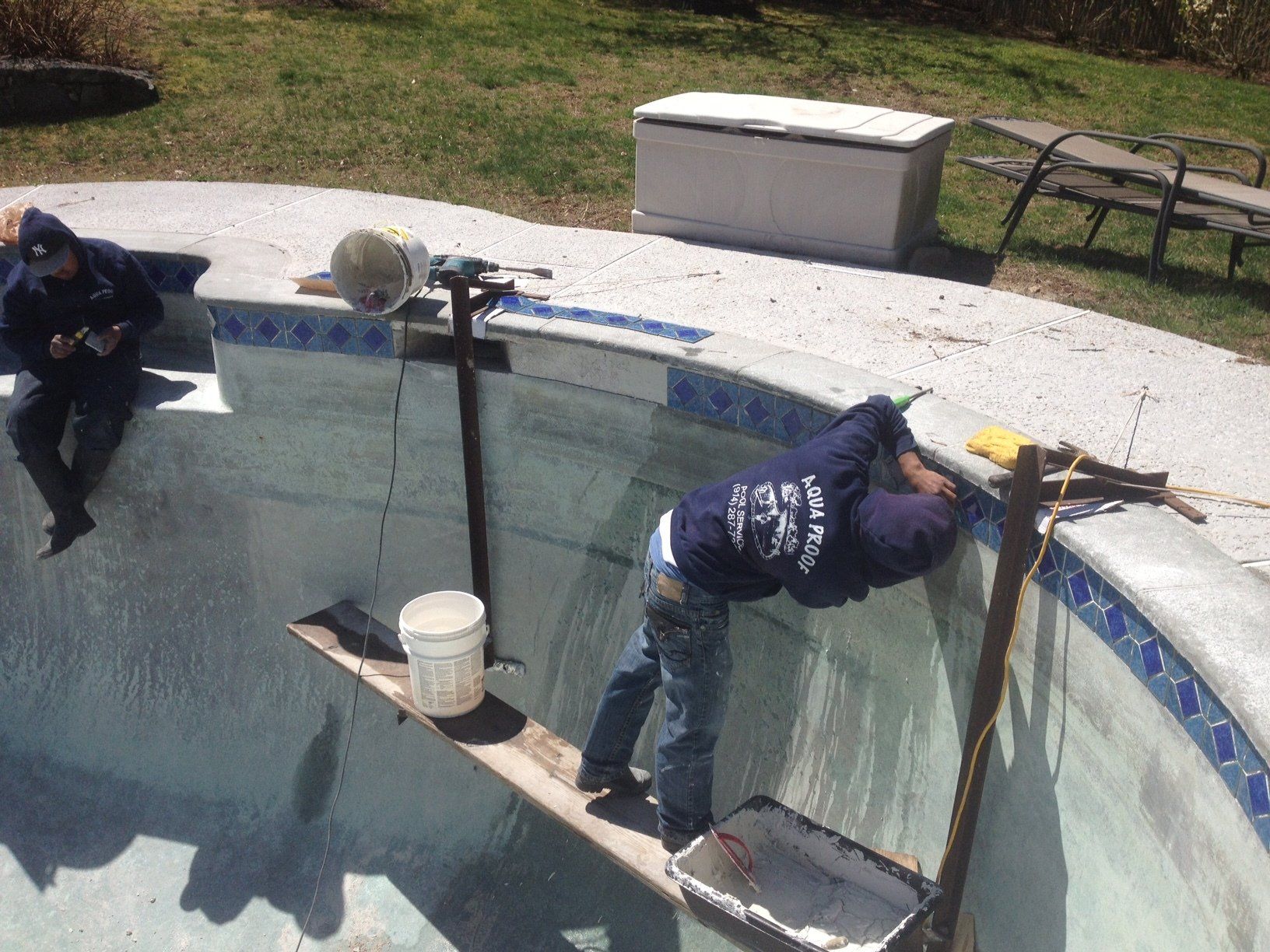 Two people repairing cracked pool walls, using tools, in a sunny outdoor setting