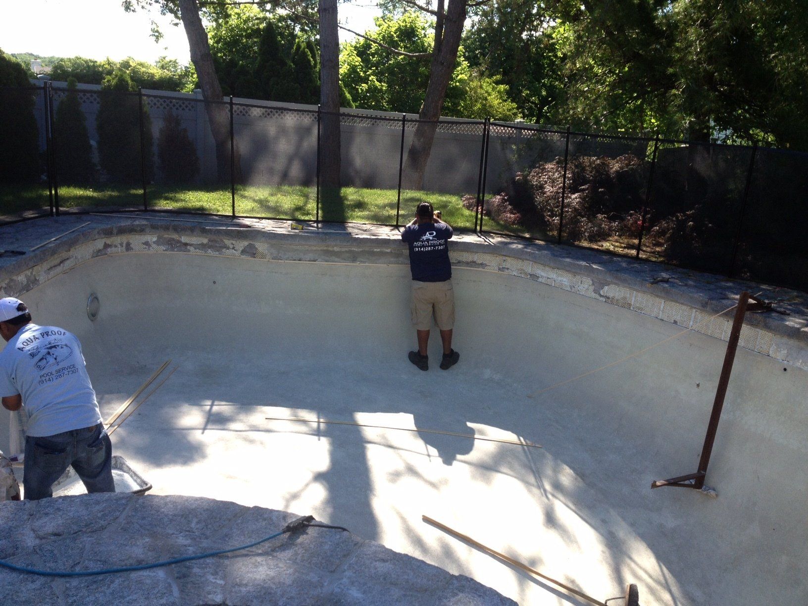 Two workers in a drained pool, one taking a photo, the other working, surrounded by fence and trees
