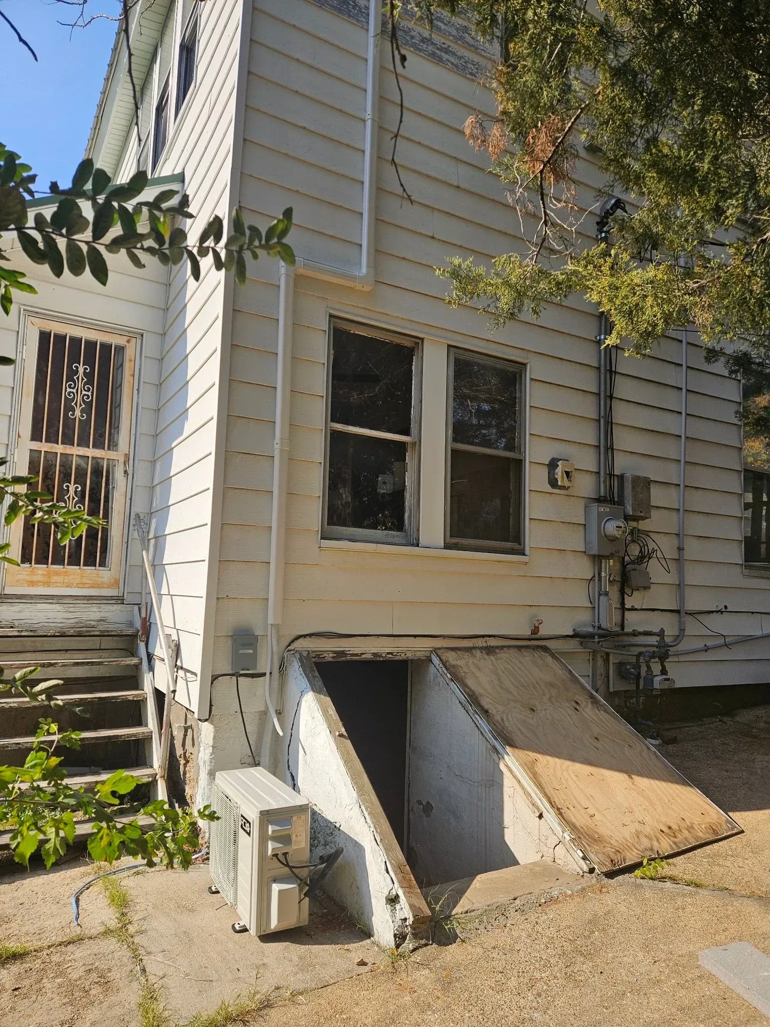 Back entrance of a two-story white house with a basement door, two windows, and air conditioning units.